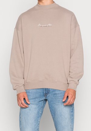 Mężczyzna w beżowej bluzie crewneck z małym białym napisem „Abercrombie New York City”, w połączeniu z jasnoniebieskimi dżinsami.