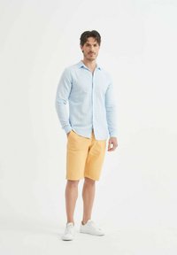 Chemise à manches longues bleu clair associée à un short jaune, avec un col standard et un design à boutons. Portée avec des baskets blanches.