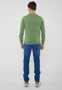 Maglione verde a maniche lunghe con scollo rotondo, abbinato a jeans in denim blu e sneakers chiare, visto da dietro.