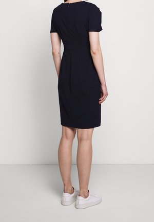 Robe bleu marine à manches courtes avec une taille cintrée, un ourlet arrivant au genou et une fermeture éclair au dos. Portée avec des baskets blanches. Texture de tissu lisse.