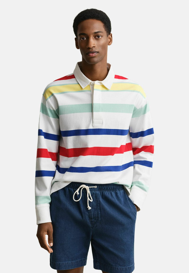 Uomo che indossa una maglia da rugby bianca a maniche lunghe con strisce orizzontali multicolori e pantaloncini da ginnastica blu navy con coulisse, in piedi davanti a uno sfondo uniforme.