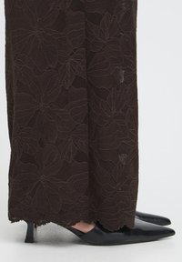 Juodos nėrinių kelnės su gėlių raštu ir banguota apačia, derinamos su juodais aukštakulniais batais su smailiu priekiu.