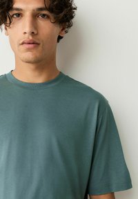 Tricou bleu din bumbac, cu guler rotund, confecționat dintr-un material moale, având mâneci scurte și o croială lejeră, cu o textură netedă și detalii de cusătură minime.