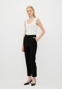 Witte geribde tanktop, op maat gemaakte zwarte broek met een zak, gedragen met glanzende zwarte pumps met een gouden accent. Model staat tegen een effen achtergrond.