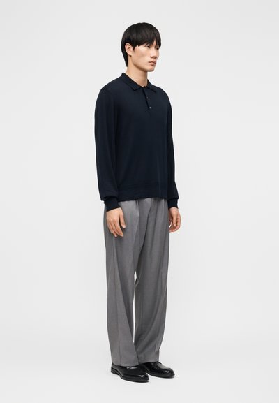 Noor mees seisab, kandes musta pika varrukaga polo särki, halli laia säärega pükse ja musti kingi ühtlase tausta ees.