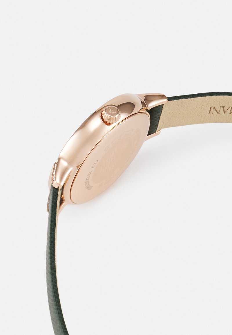 Złota różowa koperta zegarka w okrągłym kształcie z teksturowanym, zielonym skórzanym paskiem. Posiada koronę oraz grawerowane detale na tylnej części koperty.