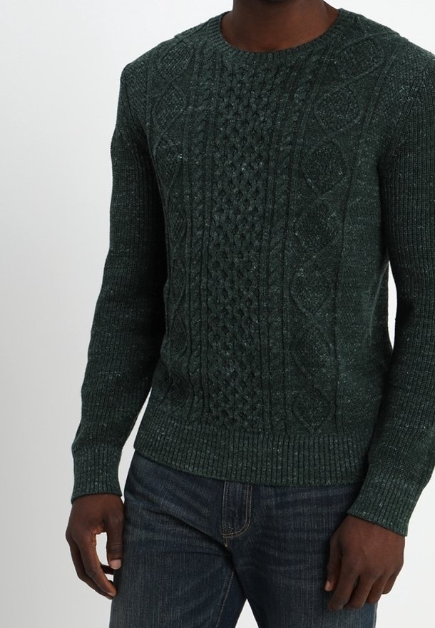 Grüner Strickpullover mit einem strukturierten Zopfmuster sowie geripptem Saum und Bündchen. Kombiniert mit dunklen Jeans.