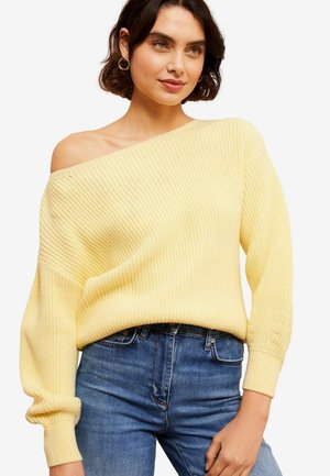 Gul strikket off-shoulder sweater med ribbet tekstur, løs pasform og lange ærmer, parret med mørkeblå jeans.