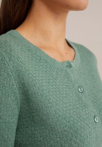 Woman wearing a green textured knit cardigan with round neckline and buttons, shown from shoulder to neck against a neutral background.
