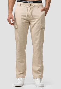 Beige cargo hlače s remenom u struku, s bočnim džepovima i glatkim, laganim materijalom. Nosene s bijelim tenisicama.