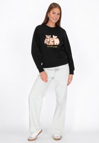 Czarna bluza z grafiką kota i żółtym napisem, zestawiona z jasnymi, luźnymi dżinsami oraz białymi trampkami.