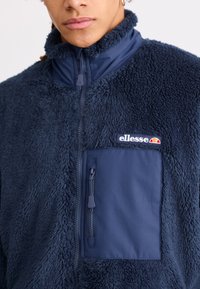 Mornarska flis jakna s visokim ovratnikom i zatvorenim džepom na prsima. Ima teksturiranu, pahuljastu površinu i oznaku s logotipom na lijevoj strani prsiju.