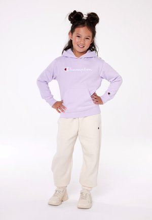 Sudadera morada con logo blanco de "Champion", bolsillo frontal. Pantalones de chándal crema con logo en la cadera, puños acanalados, combinados con zapatos claros.