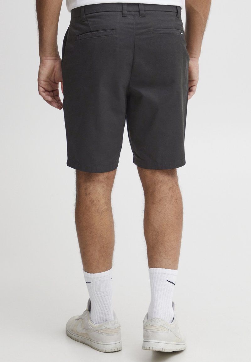 Shorts noirs en tissu léger, à coupe droite, avec deux poches arrière et une ceinture avec passants. Associés à des chaussettes et des chaussures blanches.