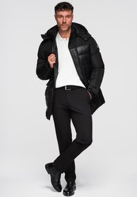 Giubbotto imbottito nero con design trapuntato, cappuccio e chiusura con zip, abbinato a una polo bianca e pantaloni neri, indossato con scarpe nere.