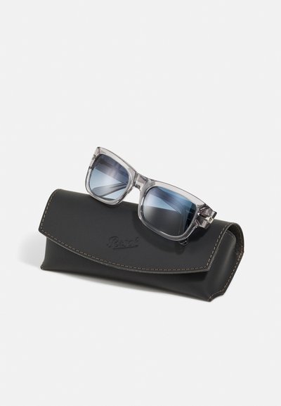 Óculos de sol com armações cinza transparentes e lentes azuis de degradê, repousando sobre um estojo de couro preto com costura castanha.