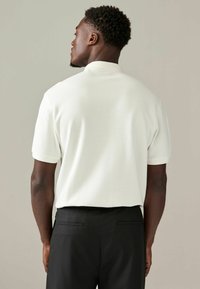 Witte poloshirt met geribbelde textuur, korte mouwen en een opgestikte kraag, gecombineerd met zwarte broek met een gladde afwerking en zakdetails.