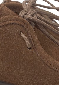 Scarpa in suede marrone con una texture liscia, forma arrotondata e lacci intrecciati attraverso piccoli occhielli per una chiusura sicura.