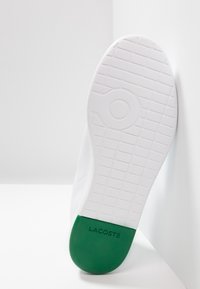 Semelle de basket blanche inclinée contre un mur, présentant un motif texturé et une section de talon verte portant l'inscription "LACOSTE".