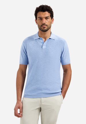 Man met een lichtblauw poloshirt met korte mouwen en beige broek, staand met één hand in de zak tegen een effen achtergrond.