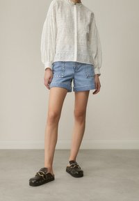 Blusa bianca in pizzo con maniche a sbuffo, abbinata a pantaloni corti di denim chiaro. Zoccoli neri con accessori dorati e dettagli borchiati completano l'outfit.