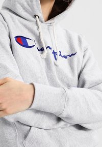 Sudadera gris con capucha, que presenta un bordado del logo de Champion en azul y rojo. Tiene un corte relajado y cordones para ajustar.