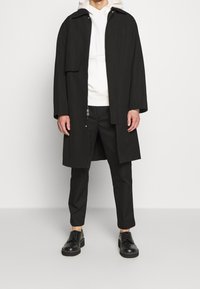 Manteau long noir avec un col montant, porté sur un sweat à capuche blanc zippé. Associé à un pantalon noir et des chaussures à lacets. Design minimaliste.