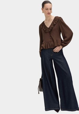 Vrouw die een bruine blouse met franjes en knopen draagt, gecombineerd met donkere jeans met wijde pijpen, en een kleine donkere handtas vasthoudt, staand tegen een effen achtergrond.