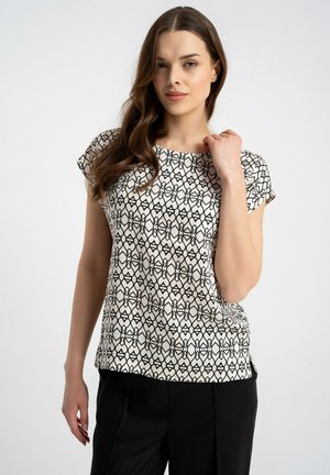 Kobieta z długimi brązowymi włosami, ubrana w czarno-biały krótki rękaw z wzorem i czarne spodnie, stojąca na tle jednolitego jasnego tła.