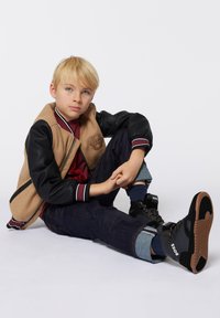 Giacca varsity marrone e nera con logo, t-shirt rossa, jeans scuri e sneakers alte nere. Materiale morbido con dettagli a righe.