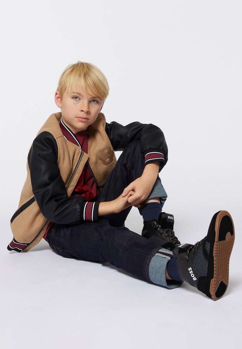 Giacca varsity marrone e nera con logo, t-shirt rossa, jeans scuri e sneakers alte nere. Materiale morbido con dettagli a righe.