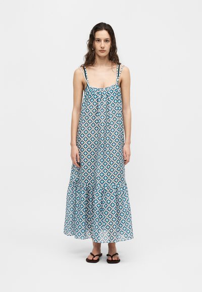Femme aux cheveux ondulés portant une robe longue à motifs géométriques bleu et blanc avec de fines bretelles et des sandales noires, debout devant un fond uni.
