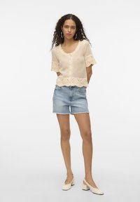 Cardigan en tricot crème avec des manches courtes à volants et une fermeture à boutons, associé à un short en denim bleu clair et des chaussures à talons bloc crème.