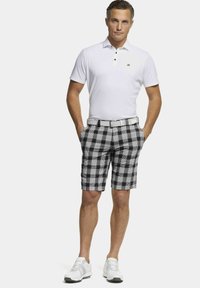 Weißes Poloshirt mit drei Knöpfen. Schwarze und weiße kariert Shorts mit Taschen und einem weißen Gürtel. Weiße Sportschuhe, im Stehen.