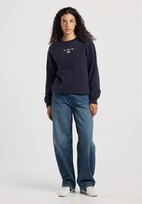 Joven con cabello rizado que lleva un sudadera azul marino con el texto "New York City", jeans azules holgados y zapatillas blancas, de pie frente a un fondo blanco.