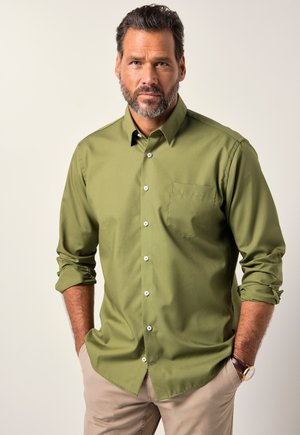 Camicia verde oliva con bottoni, maniche lunghe, tasca sul petto a sinistra, bottoni bianchi e una texture liscia. Abbinata a pantaloni leggeri beige.