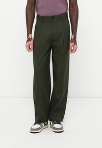 Pantalones plisados verdes con un corte recto, hechos de una tela suave. Llevados con una camisa morada y zapatillas negras y blancas.