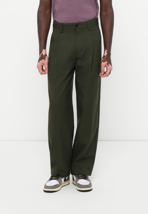 Green pleated trousers with a straight fit, made from a smooth fabric. Worn with a purple shirt and black and white sneakers.
