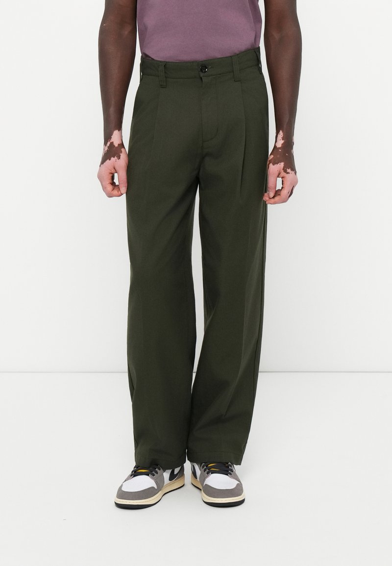 Pantalones plisados verdes con un corte recto, hechos de una tela suave. Llevados con una camisa morada y zapatillas negras y blancas.