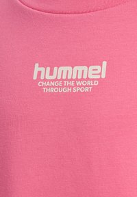Pink stof med hvid tekst "hummel SKIFT VERDEN GENNEM SPORT" centreret nær halsudskæringen.