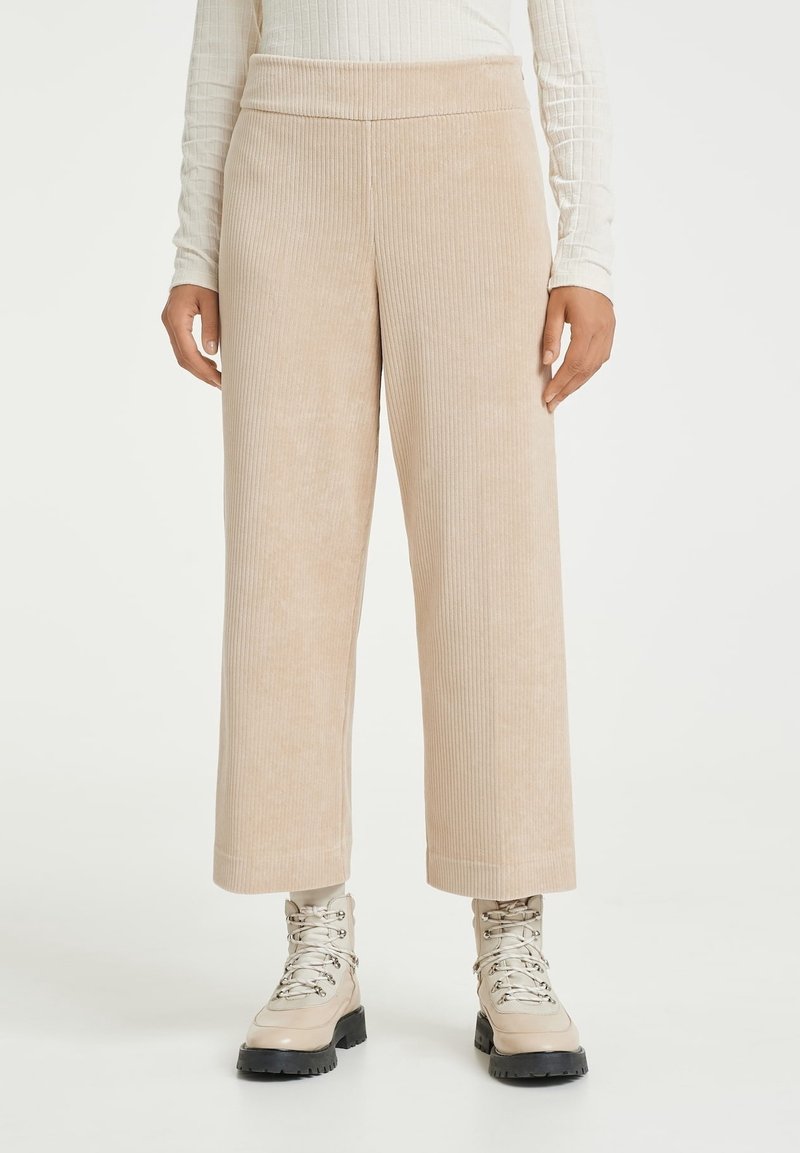 Pantalon large en velours côtelé beige porté avec des bottines beiges claires à lacets sur une personne portant un haut crème côtelé à manches longues.