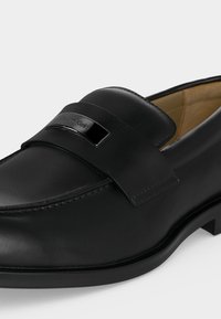 Ανδρικό μοκασίνι από μαύρο δέρμα με λεία υφή, διακοσμητικό λουράκι και μεταλλική πινακίδα με το λογότυπο Calvin Klein στο πάνω μέρος.