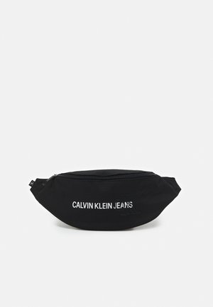 Czarna nerka Calvin Klein Jeans z zamkiem błyskawicznym i białym nadrukowanym logo z przodu, na jednolitym jasnym tle.