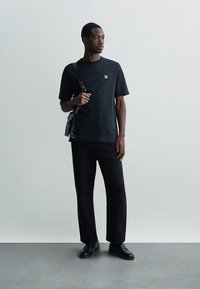 Mann trägt ein schwarzes T-Shirt mit kleinem Emblem, schwarze Hosen, schwarze Schuhe und hält eine schwarze Umhängetasche vor einem schlichten hellen Hintergrund.