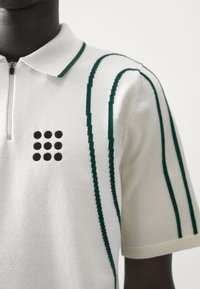Polo shirt in cream knit fabric featuring a zip collar, green stripes, and a black dot pattern on the chest. Short sleeves with ribbed trim.