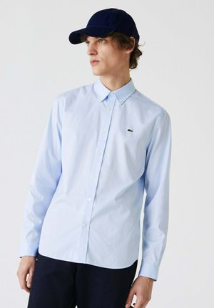 Hellblaues Button-Up-Hemd mit langen Ärmeln und klassischem Kragen, mit einem kleinen grünen Logo. Wird mit einer marineblauen Baseballkappe getragen.