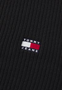 Tissu en tricot noir côtelé avec un patch logo brodé rectangulaire portant le texte "TOMMY JEANS" et des blocs de couleurs rouge, blanc et bleu.
