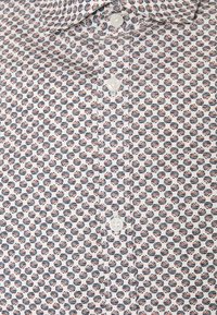 Camisa blanca con un patrón repetitivo de semicirculos en azul marino y rojo. Cierre de botón con botones de plástico blanco. Textura de tela suave.