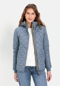 Quilted blauwe jas met capuchon, voorzien van een ritssluiting aan de voorkant, een gegerande textuur en ribgebreide manchetten. Gepaard met een witte top en jeans.