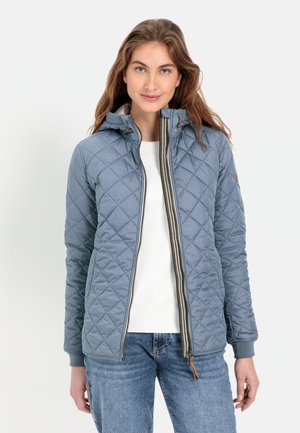 Quilted blauwe jas met capuchon, voorzien van een ritssluiting aan de voorkant, een gegerande textuur en ribgebreide manchetten. Gepaard met een witte top en jeans.